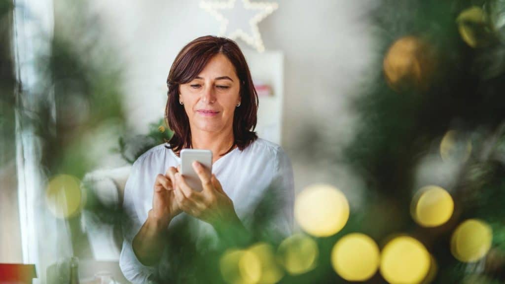 A woman using a smartphone indoors, framed by soft holiday lights and greenery.