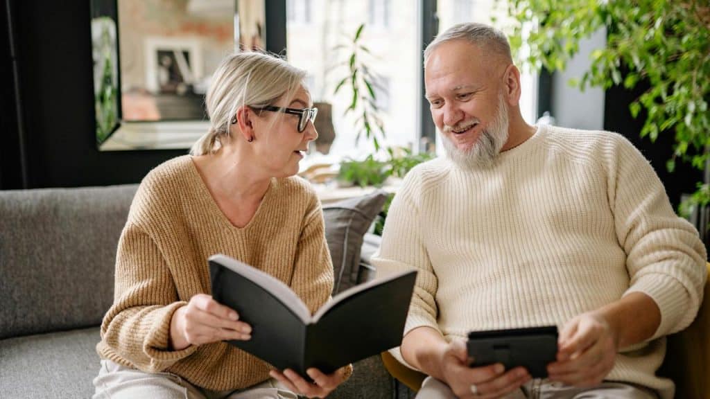 A senior couple sitting on a couch, smiling while looking at a book together.