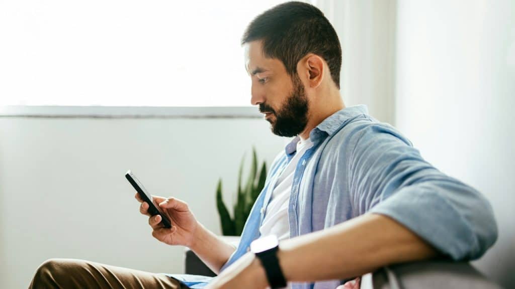 A bearded man sitting on a couch and using his phone.