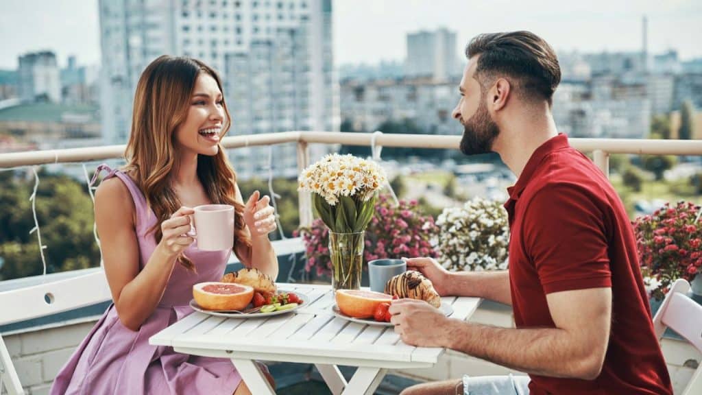 A man and woman sit at an outdoor table with breakfast, talking and smiling together.