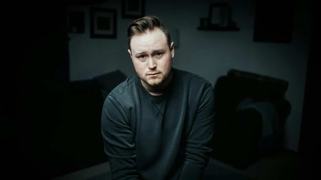 A man in a dark sweatshirt sits in a dimly lit room looking directly at the camera.