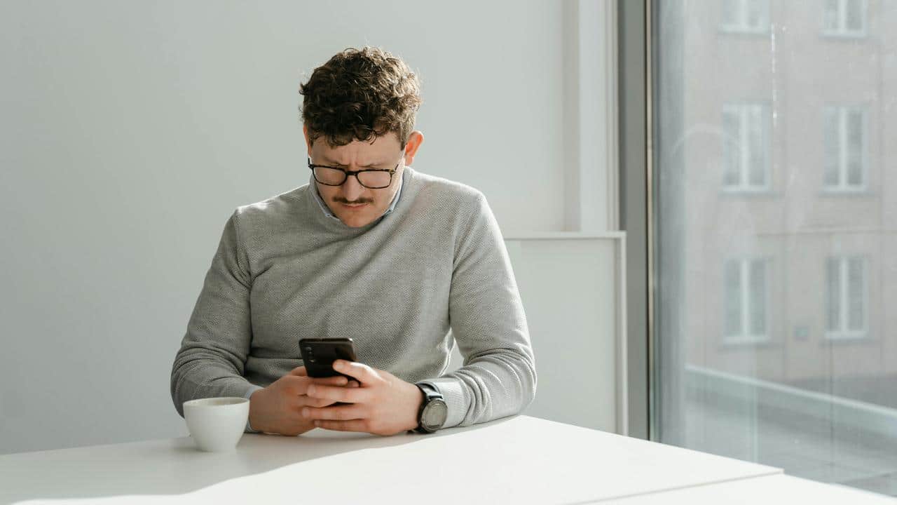 A man wearing glasses sits at a table looking at his phone with a cup nearby.