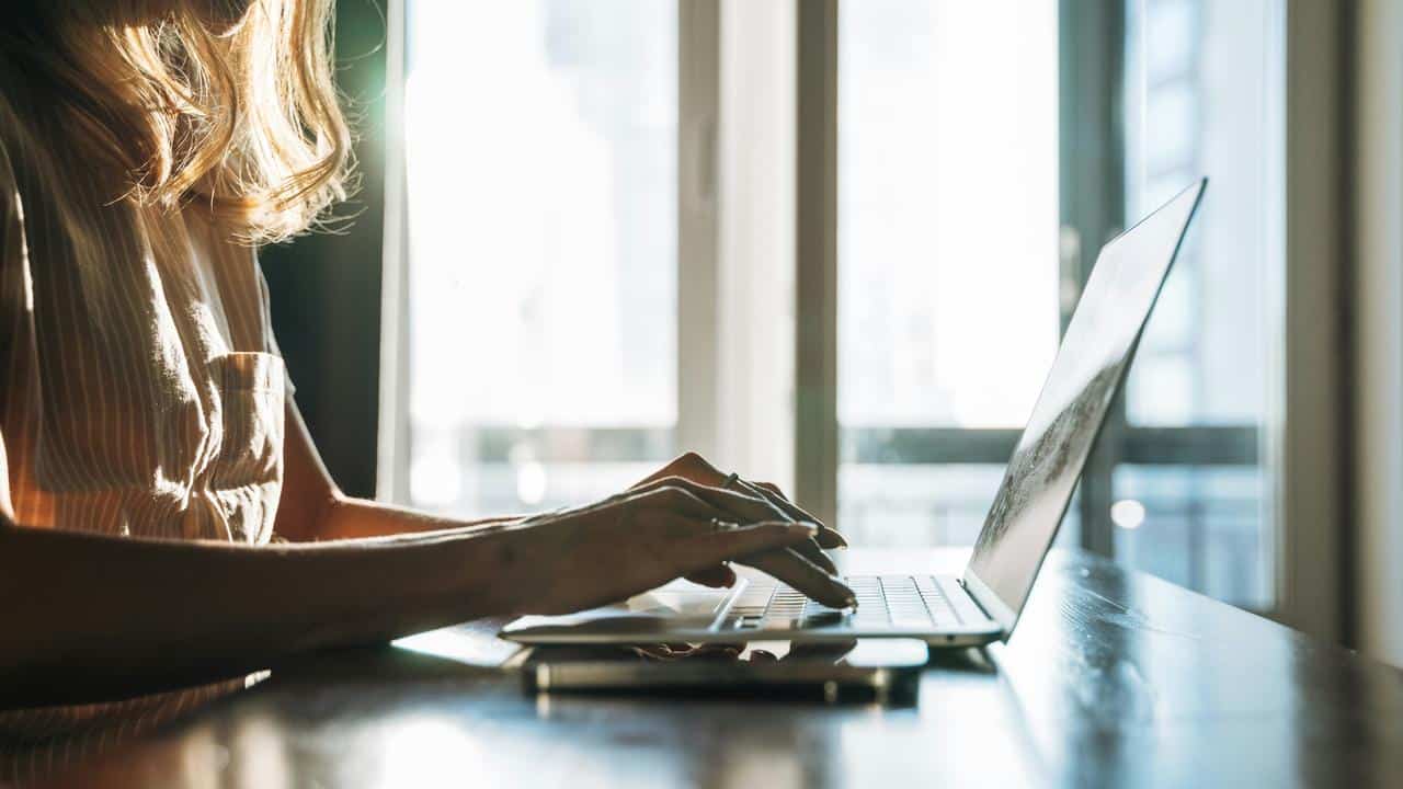 A person typing on a laptop at a desk near a window.