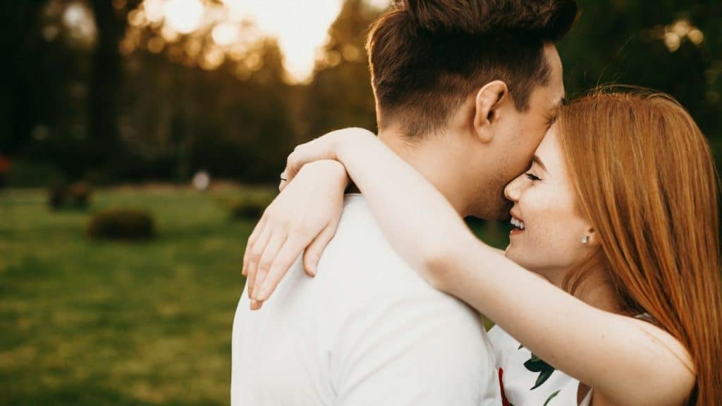 A woman with red hair smiles as she embraces a man in a grassy field.