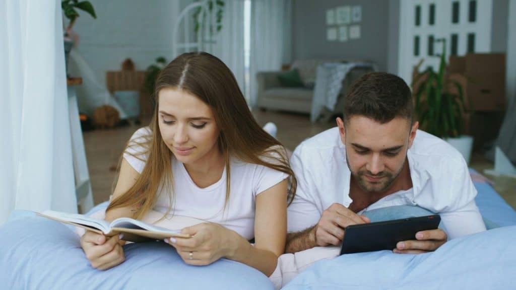A man and woman lie on a bed; she reads a book while he uses a tablet.