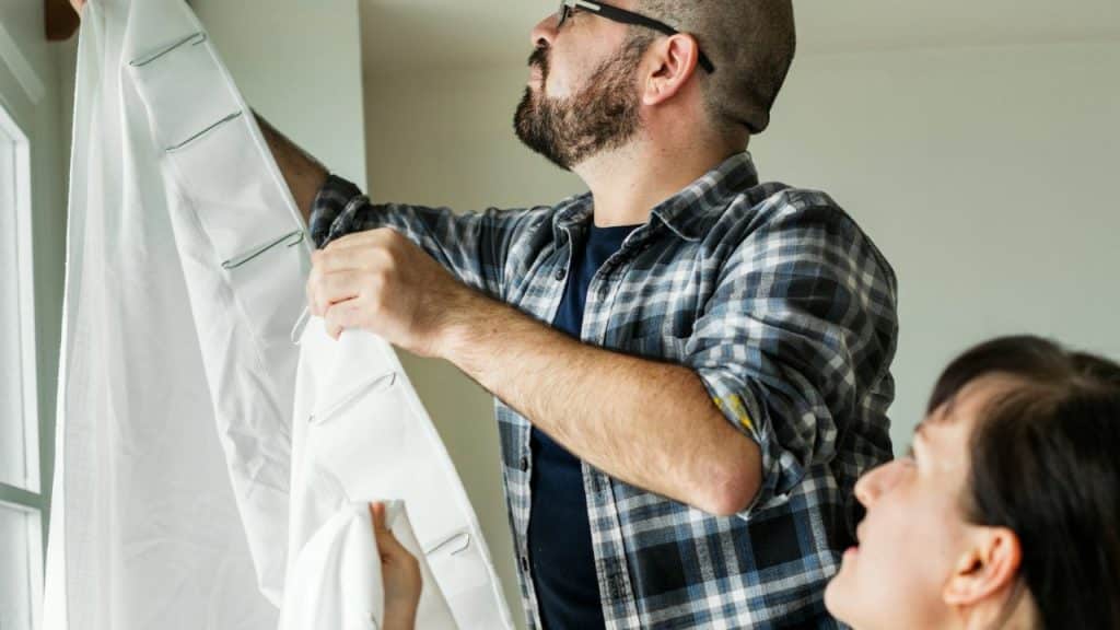 A man in a plaid shirt hangs white curtains while a woman looks on.