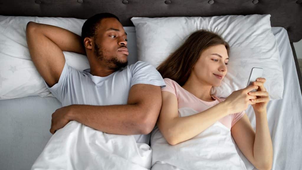 A man and woman lying in bed, with the man looking suspiciously at the woman who is focused on her phone.