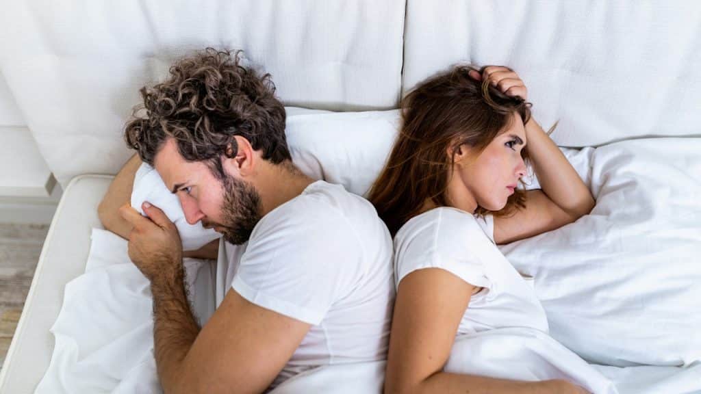 A couple lying in bed, facing away from each other with distressed expressions.
