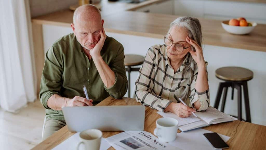 A serious older couple works at a kitchen table, reviewing bills, writing notes and using a laptop.
