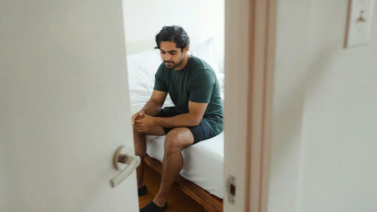 Man sitting on the edge of a bed, viewed through a partially open doorway.