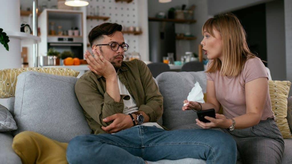 A man sits looking bored on a couch while a woman talks animatedly holding a tissue.