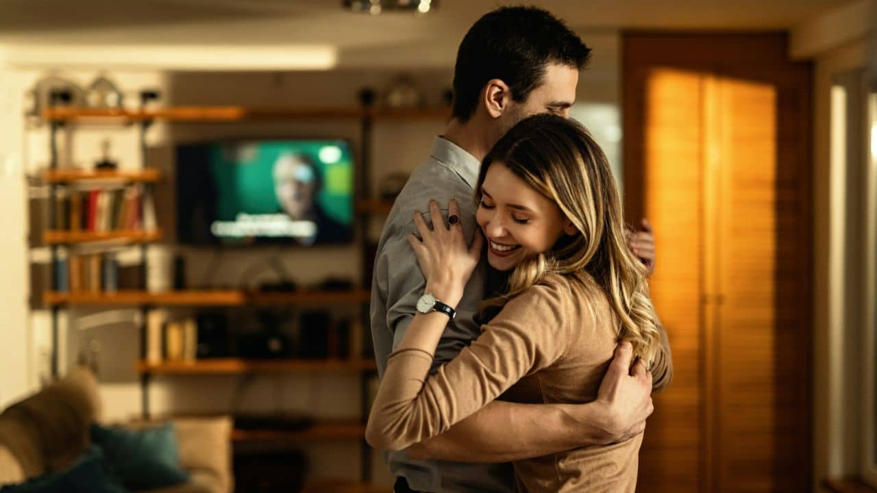 A man and woman embrace and smile in a dimly lit living room with a television in the background.