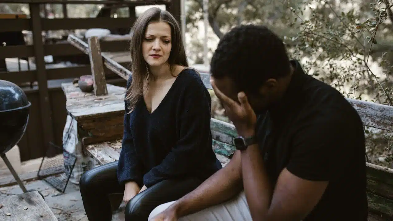 A woman sits next to a man who is holding his head in his hands outdoors.