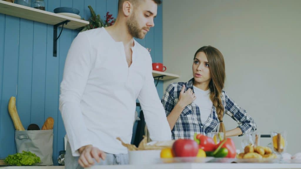 Woman in a plaid shirt looking seriously at a man in a white shirt in a kitchen.