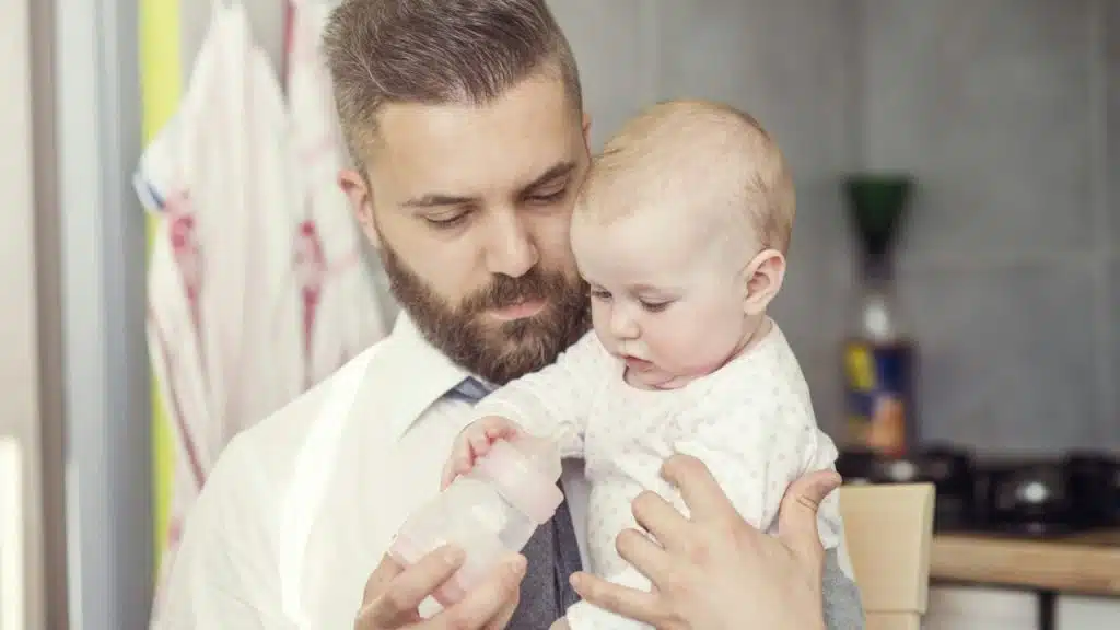 Bearded man in white shirt and tie holding and feeding a baby a bottle.
