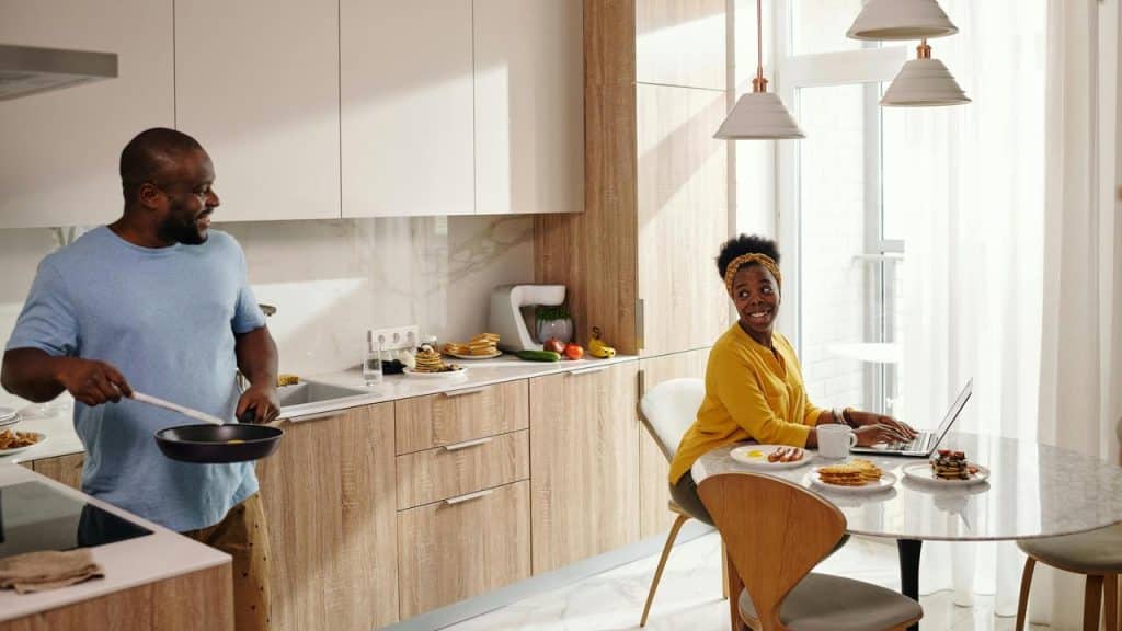 A man cooking breakfast while a woman smiles at him from the kitchen table.