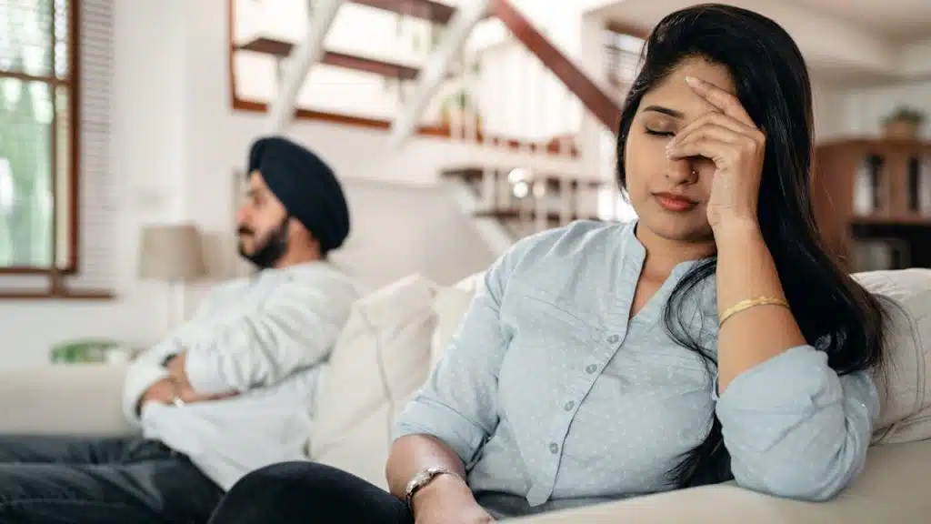 A distressed woman is sitting on a couch with her hand on her forehead, while a man wearing a turban sits turned away from her
