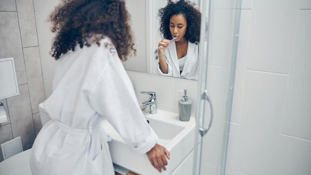 A woman in a bathrobe brushes her teeth while looking in the bathroom mirror.