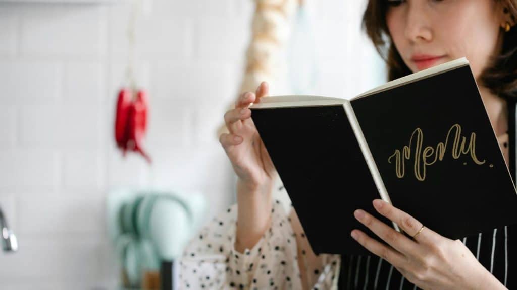 A woman reading a black menu book in a kitchen.