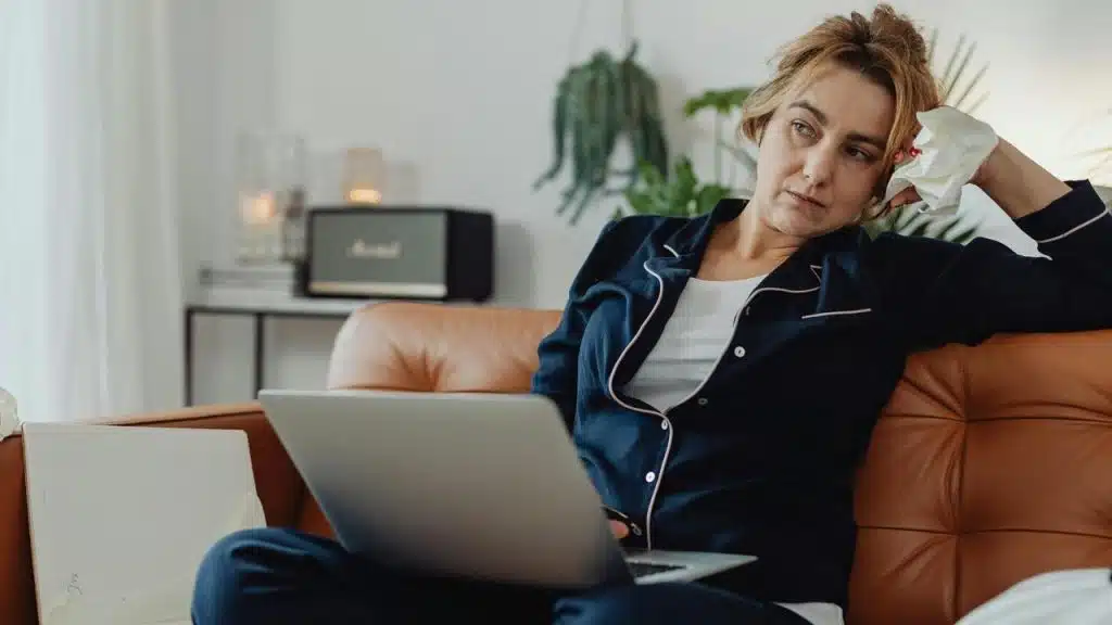 A woman sitting on a couch with a laptop on her lap holding a tissue and looking tired.