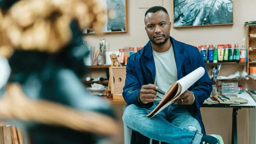 A man in a blue coat sits in an art studio, holding a sketchbook and looking forward.
