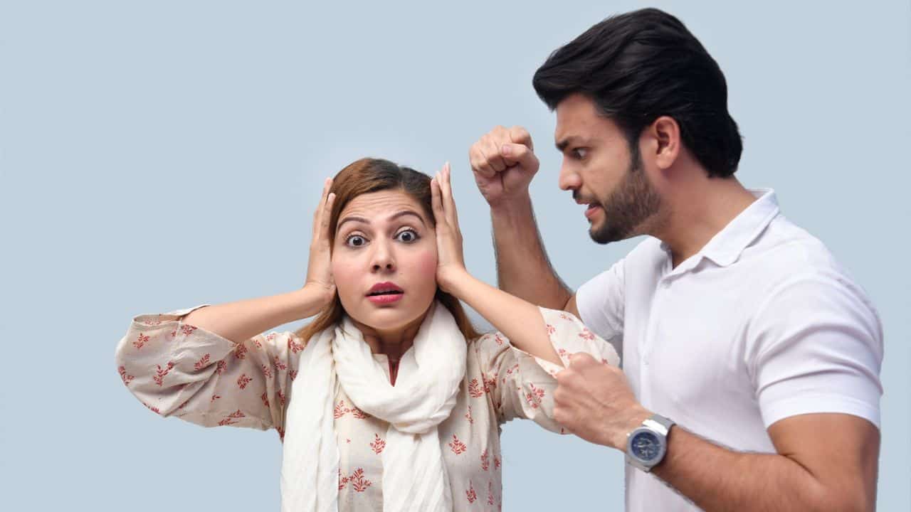 A man with a raised fist shouts at a terrified woman covering her ears in front of a blue background.