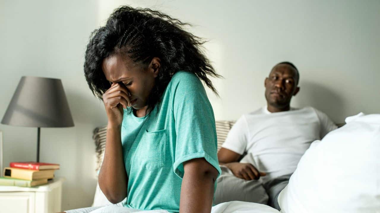 A woman with her face in her hands sits on a bed, a man looks forward behind her.