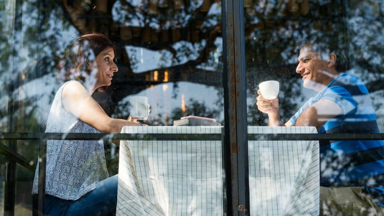 Couple enjoying coffee and conversation at a table viewed through a window outdoors.