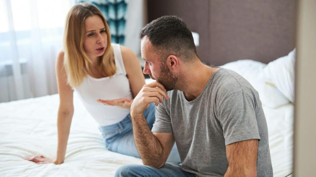 Man sits on a bed looking upset while a woman gestures and speaks angrily behind him.
