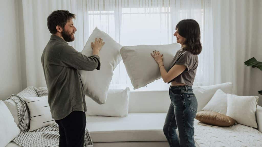 Smiling couple holding large white pillows in a living room, facing each other.