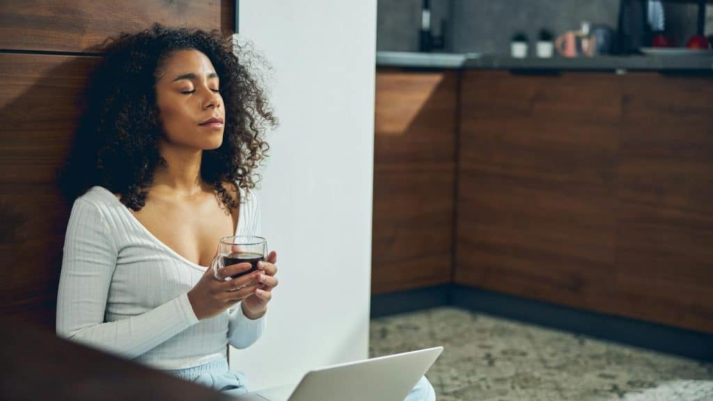 A woman sits on the floor with her eyes closed, holding a cup and relaxing beside a laptop.