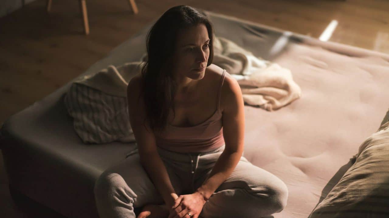 Woman sitting alone and looking distressed on a bed in a dimly lit room.
