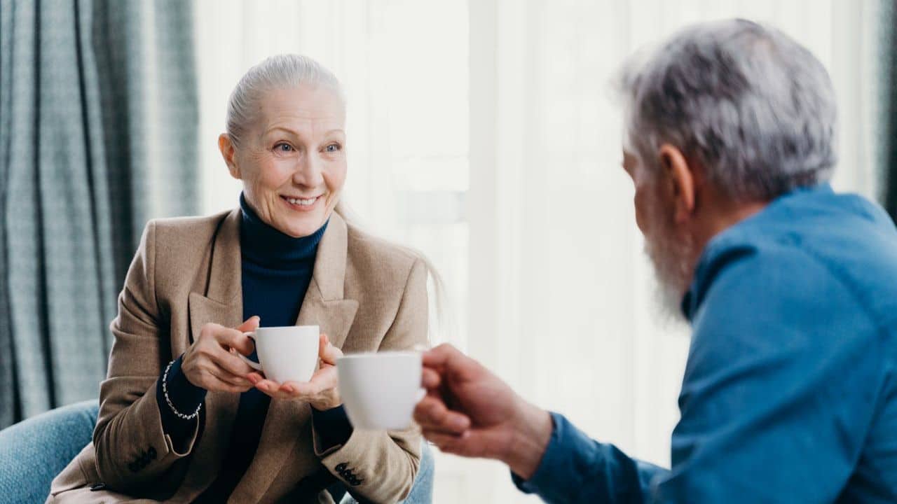 Smiling older woman holding a coffee cup, looking at a man across from her.