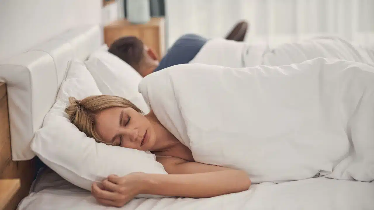 A blonde woman is sleeping on her back while a man sleeps with his back to her in the same bed.