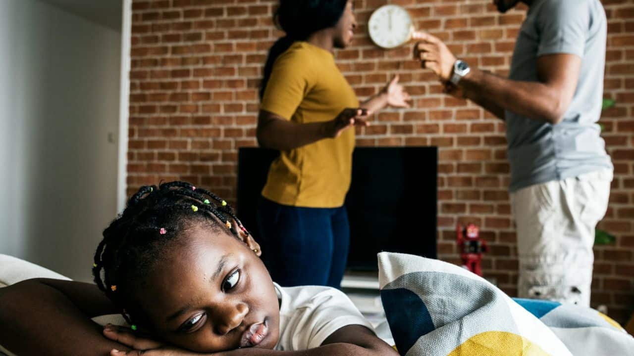 Young girl looking sad and worried while parents argue in the background.