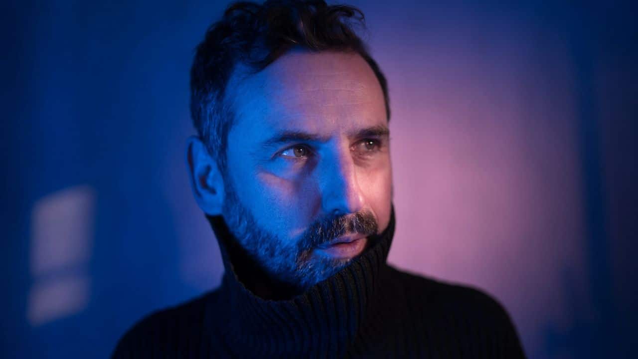 A middle-aged man in a dark turtleneck is illuminated by blue and pink light.