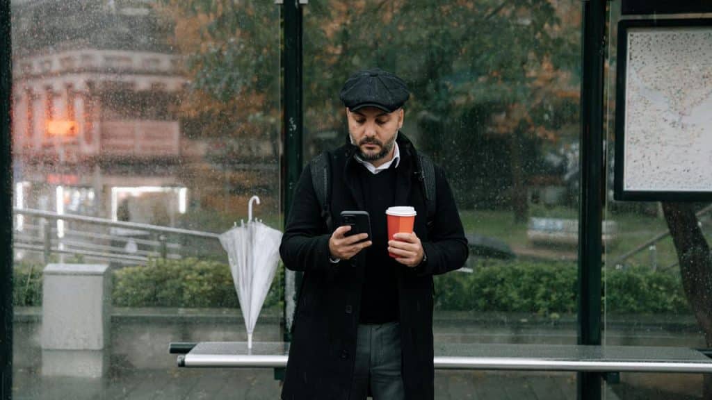 A man standing at a bus stop holding a coffee cup and using his smartphone