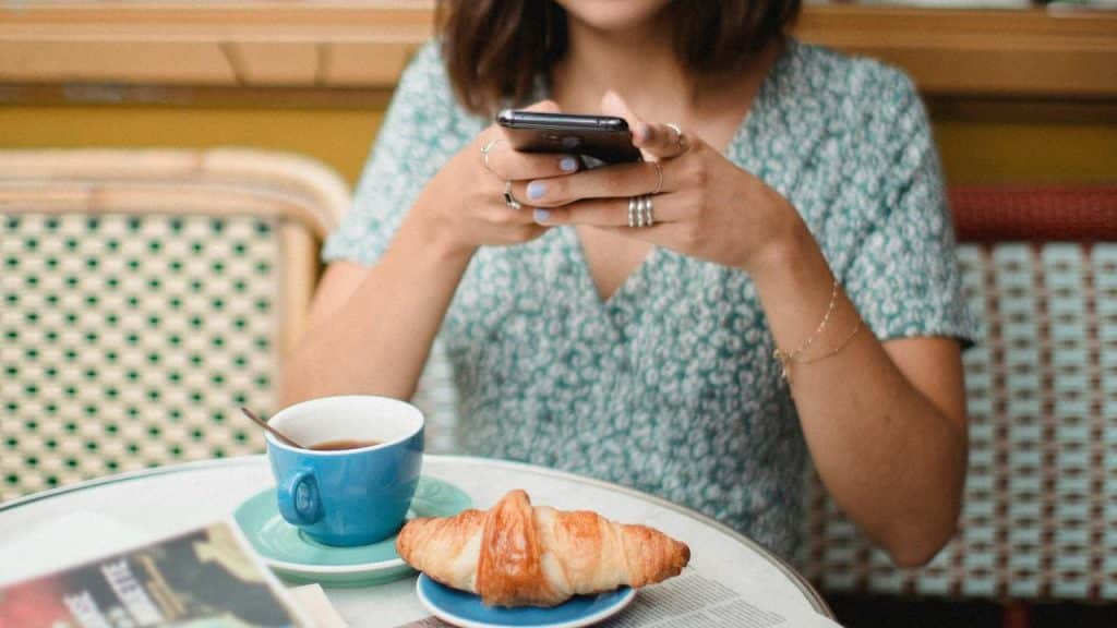 A person sitting at a café table using a phone with coffee and a croissant.