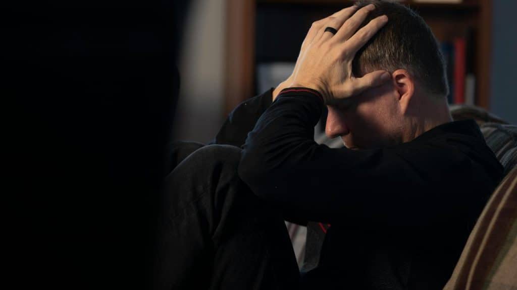 A person sitting on a couch with their hands on their head, appearing distressed.
