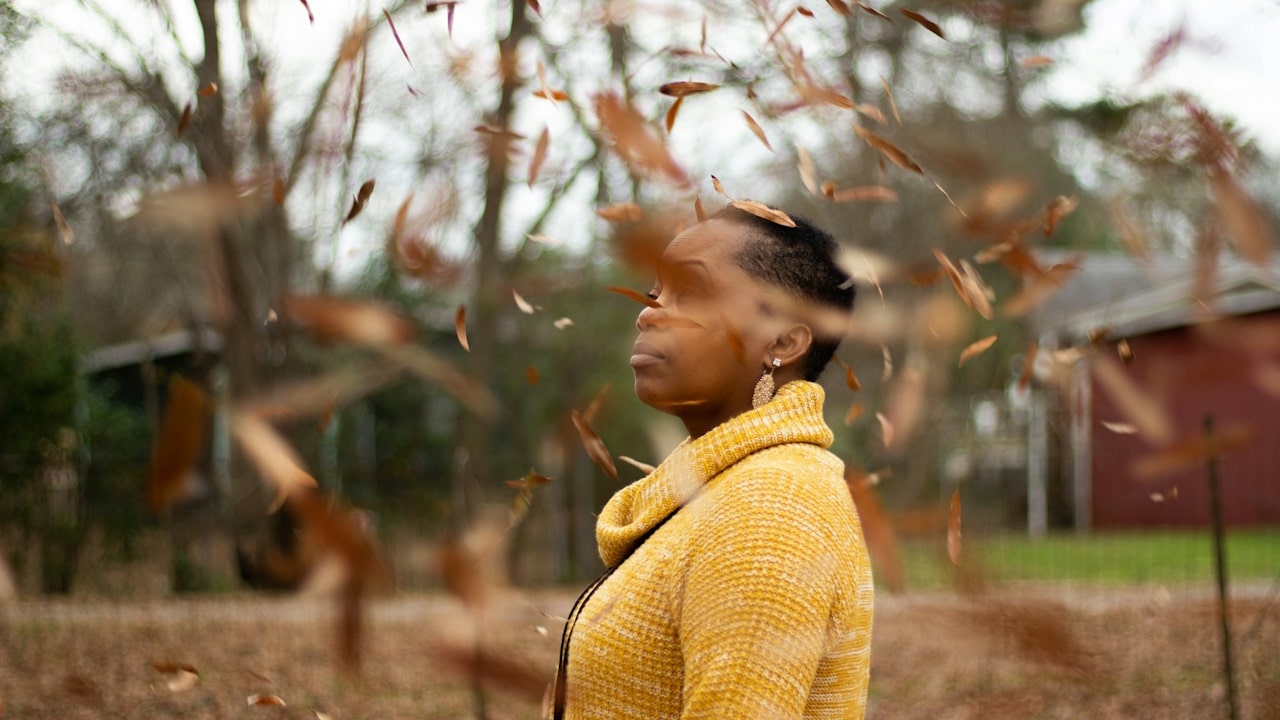 A woman in a yellow knit sweater standing near brown trees during the daytime.