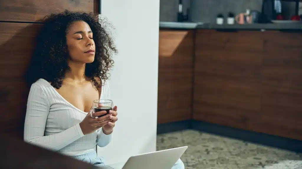 A woman sits on the floor with her eyes closed, holding a cup and relaxing beside a laptop.