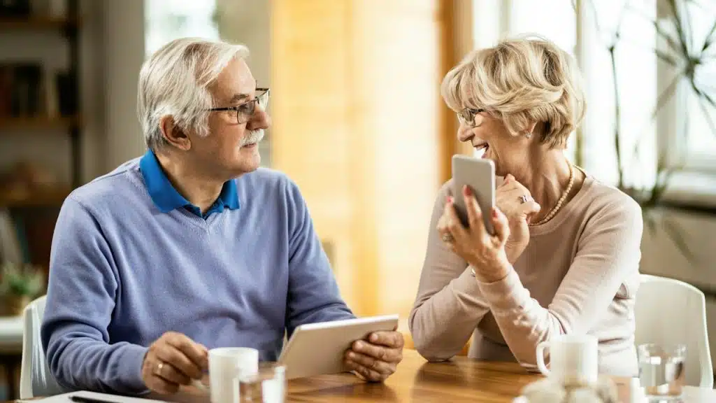 A mature couple talking as the man has coffee and his wife smiles at him while using her phone.