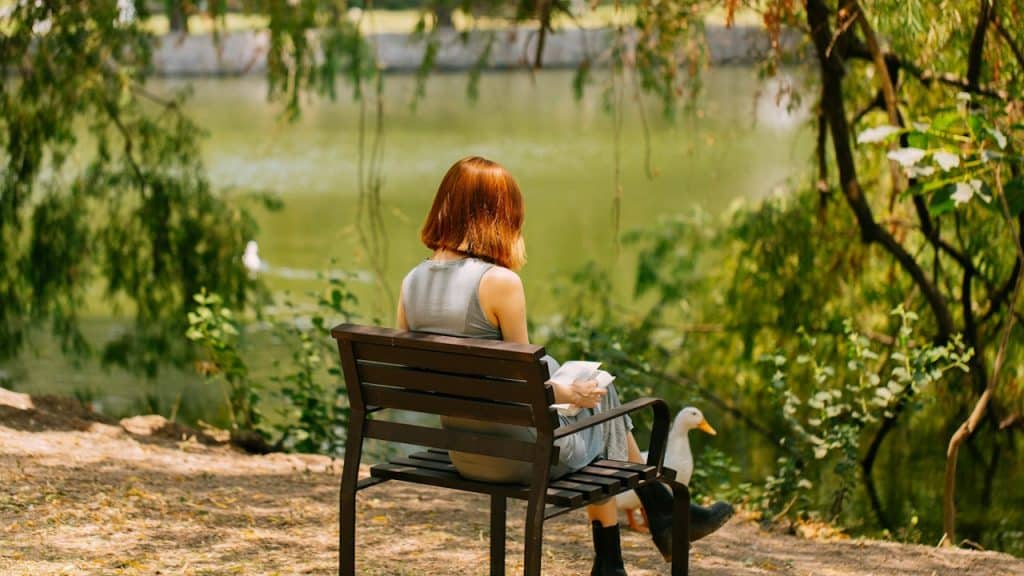 A woman sitting on a bench by a lake and reading a book.