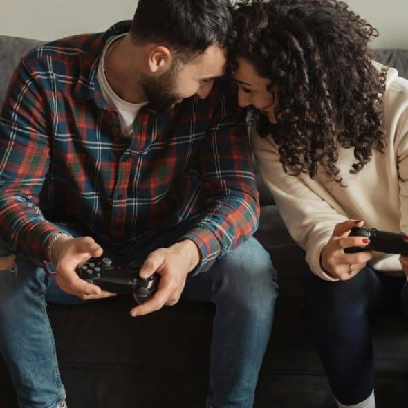 A couple sitting on a couch smiling while playing video games with controllers.