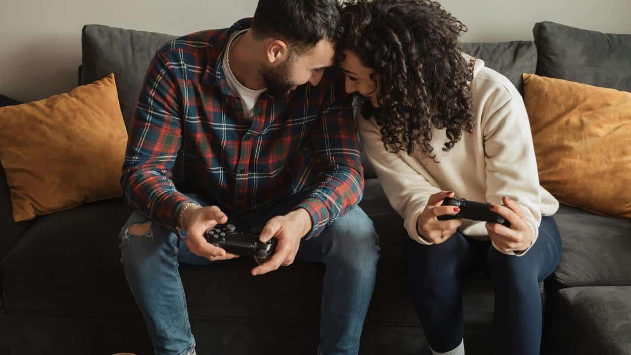 A couple sitting on a couch smiling while playing video games with controllers.