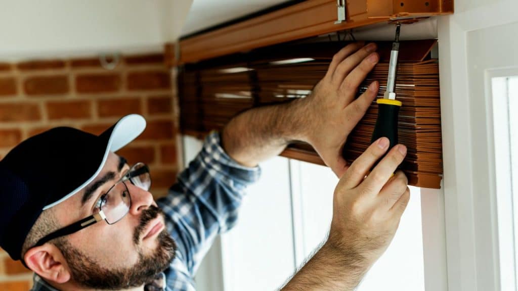 A man in a cap and glasses installs wooden blinds with a screwdriver.