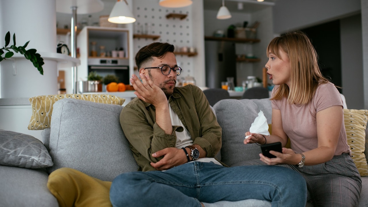 A woman speaking intently to a man who is looking away, as they sit on a couch in a living room.
