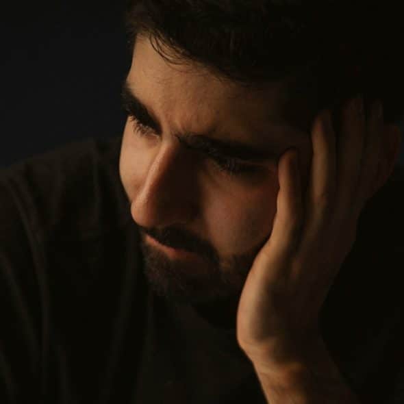 A bearded man looking upset and holding his face with his hand.
