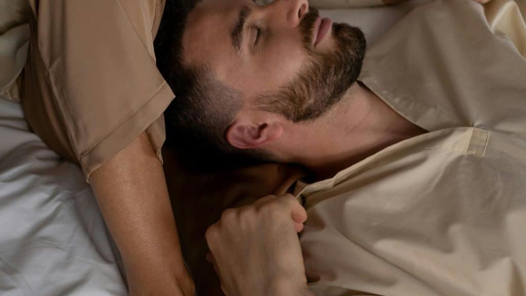 A bearded man resting on a bed with his eyes closed, holding a bedsheet.