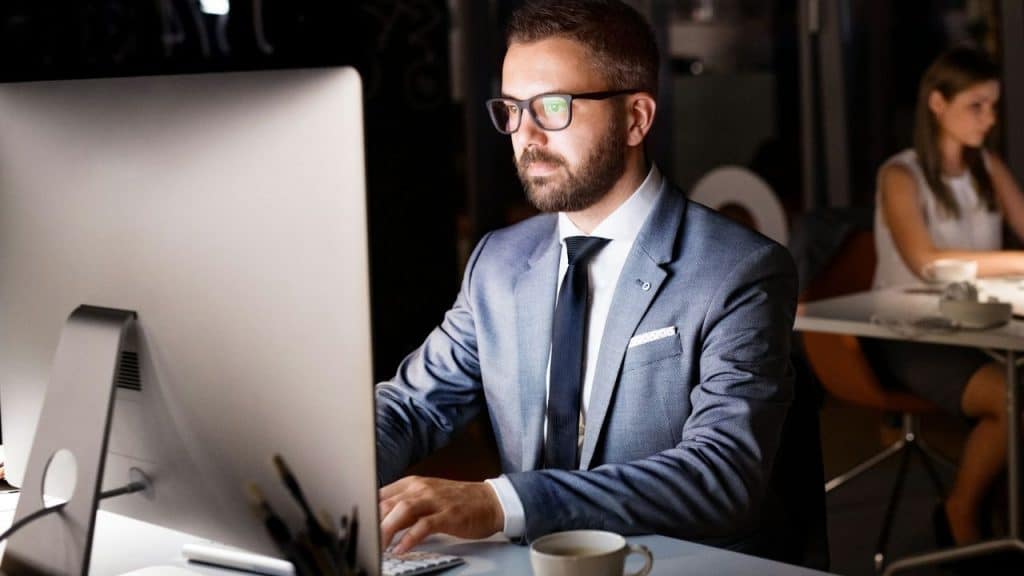 Man in a suit and glasses working late on a computer in a dark office.
